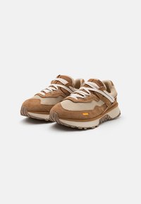 Scarpe atletiche in suede marrone e rete beige con lacci bianchi piatti, suole texturizzate e un piccolo accento arancione vicino alla suola.