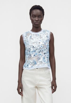 Femme portant un haut transparent sans manches orné de sequins bleus et argentés, associé à un pantalon blanc taille haute, debout devant un fond uni.