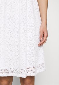 White lace dress with floral pattern, a flared shape, and scalloped hem. The texture is delicate, complemented by a subtle transparency.