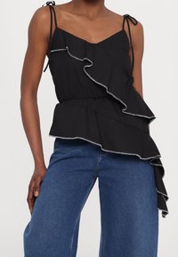 Schwarzes ärmelloses Top mit asymmetrischen, fransigen Lagen und Bindebändern, kombiniert mit blauen Denim-Jeans an einer stehenden Person.