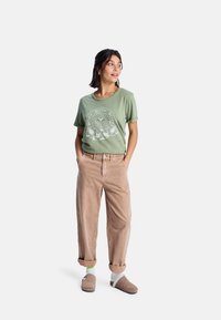 T-shirt vert en coton avec imprimé floral blanc et logo, associé à un pantalon en velours côtelé beige, avec les poignets retroussés, et des chaussons gris.