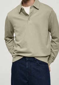 Beżowa koszula polo z długim rękawem, z kołnierzykiem i plastronem na trzy guziki, w połączeniu z ciemnymi dżinsami. Gładka tekstura, luźny krój.