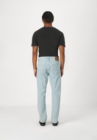 Jeans de mezclilla azul claro con un ajuste recto, que presentan un parche de cuero marrón en la parte trasera de la cintura y un diseño de cinco bolsillos. Camisa negra de manga corta.