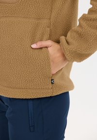 Ruskea fleece-pullovetti, jossa on teksturoitu pinta, edessä tasku ja pieni logotagi. Yhdistetty tummansinisien housujen kanssa.