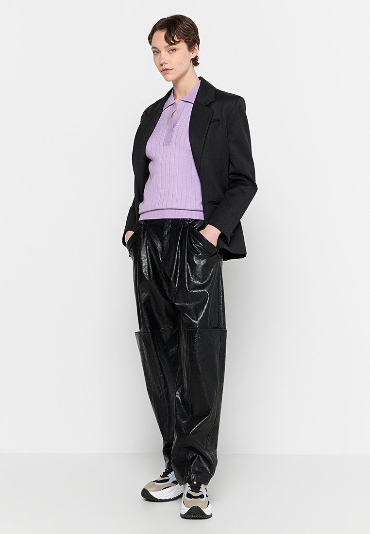 Schwarzer Blazer über einem lavendelfarbenen gerippten Polo, kombiniert mit glänzenden schwarzen baggy Hosen und mehrfarbigen Turnschuhen. Minimale Accessoires, Hände in den Taschen.