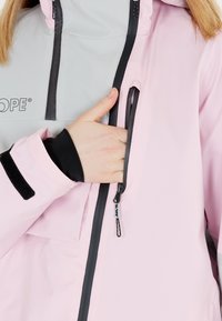 Ljust rosa vattentät jacka med grå accenter, framfickor med dragkedja, elastiska ärmslut, logga på bröstet och en säker dragkedjepull.