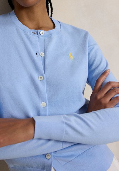 Person mit geflochtenem Haar trägt hellblauen, mit Knöpfen versehenen Cardigan mit gelbem gesticktem Logo, die Arme vor neutralem Hintergrund verschränkt.