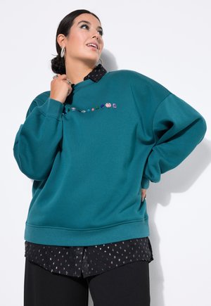 Felpa oversize color teal con girocollo, caratterizzata da decorazioni colorate di perline sul petto. Indossata sopra una maglietta nera con un motivo.