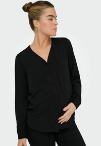 Haut noir à manches longues avec décolleté en V et fermeture par boutons sur le devant. Présente un tissu texturé et une coupe légèrement ample, adaptée à la maternité.