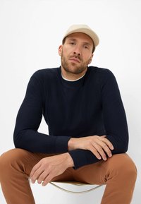 Marineblauer Strickpullover mit langen Ärmeln, rundem Halsausschnitt und gerippten Bündchen, kombiniert mit hellbraunen Hosen. Die Mütze ist beige.