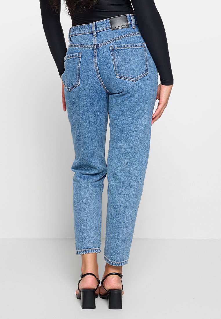 Jeans z wysokim stanem w niebieskim odcieniu, o zwężanym kroju, z pięcioma kieszeniami i subtelnym przeszyciem, połączone z czarnymi sandałami na obcasie z paskami.