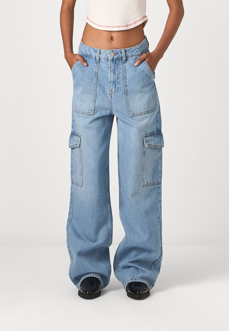 Femme portant un jean cargo large bleu clair avec grandes poches avant et latérales, mains dans les poches, avec des chaussures noires et un haut court.