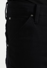 Černé džínové kalhoty mají hladkou texturu, klasický střih, šikmé kapsy a kovové knoflíky, což zdůrazňuje minimalistický design.