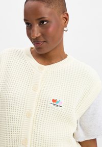 Crèmekleurige gebreide cardigan met een textuurpatroon, voorzien van een knoopsluiting aan de voorkant en een klein merkembleem in de kleuren rood, blauw en roze.
