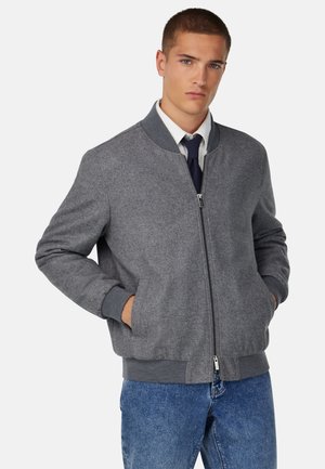 Graue Bomberjacke mit Reißverschluss, gerippten Bündchen und Kragen. Weiche Textur und zwei Seitentaschen. Über einem Hemd mit Kragen und einer Krawatte getragen.