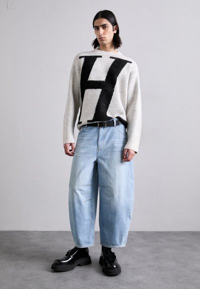 Ανοιχτό γκρι υπερμεγέθη πουλόβερ με μαύρο γραφικό 'H', συνδυασμένο με φαρδιά ανοιχτό μπλε τζιν και χοντρές μαύρες γαλότσες με υφή στη σόλα.