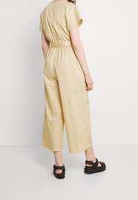 Gul mönstrad jumpsuit med korta ärmar, insvängd midja och vida byxor. Bärs med svarta platåsandaler som visar en tjock sula.