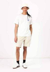 Camiseta de algodón blanca con logo azul, pantalones cortos beige y zapatillas blancas con suelas marrones. Un sombrero tipo bucket crema completa el conjunto.