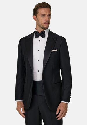 Un homme vêtu d’un smoking noir, d’une chemise blanche, d’un nœud papillon noir et d’une pochette blanche se tient devant un fond clair uni.