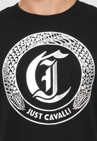 Camisa negra con un diseño de serpiente circular plateada que rodea una "J" y "L" estilizadas, con el texto "Just Cavalli" debajo.