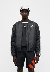 Zwarte bomberjack met witte strepen, gedragen over een zwart shirt, gecombineerd met zwarte shorts met rode accenten. De stof lijkt glad.