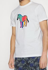 Biała bawełniana koszulka z kolorową grafiką konia z różnymi paskami w żywych kolorach. Okrągły dekolt i krótkie rękawy.