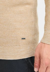 Suéter de punto beige con tela texturizada, puños y dobladillo acanalados, que presenta una pequeña etiqueta de marca gris cerca del fondo.