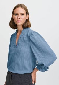 Blusa azzurra chiara con maniche a sbuffo, tessuto texturizzato e scollatura ondulata. Presenta un sottile motivo a righe verticali.
