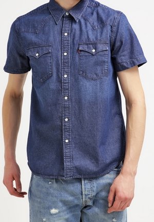 Mężczyzna ubrany w niebieską, krótki rękaw, zapinaną na guziki koszulę dżinsową z kieszeniami na piersi oraz jasnoniebieskie, podarte dżinsy na tle białego tła.