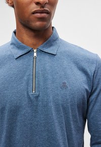 Sinise kraega poollukuga särg, valmistatud pehmest kangast. Vasakul rinnal on peen logo tikand ja lihtne disain.