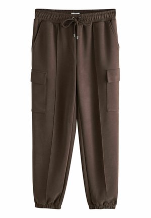 Woman with long wavy hair walking past a stone wall wearing dark loose-fitting shirt, cargo pants, flip-flops, and holding a small brown handbag.