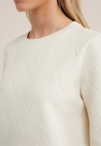 Nainen, joka pukeutuu kermaisen väriseen, tekstuuriltaan moneen muotoon ja pyöreäkaulaiseen neulepaitaan, näkyy kaulasta ylöspäin.