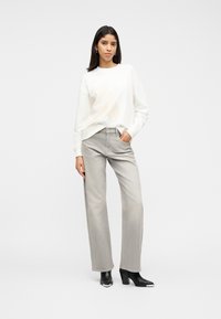 Γυναίκα που φοράει λευκό πουλόβερ με σχέδιο αλόγου, ανοιχτό γκρι τζιν φαρδιάς γραμμής και μαύρες μπότες με τακούνι, στέκεται μπροστά σε απλό φόντο.