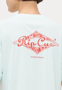 Ljust blå t-shirt med ett stort rödblommigt mönsterlogotyp på baksidan, som visar "Rip Curl" och "Surfing Company" text nedanför.