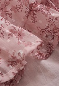 Ljusrosa tyg med ett blommönster i mörkare rosa. Tyget har en mjuk, skrynklig textur och fina volanger längs kanterna.