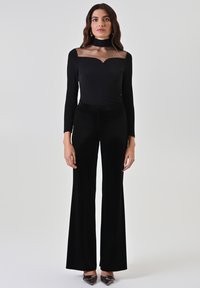 Body nero a maniche lunghe con scollo in rete trasparente e pantaloni svasati in velluto, completati da tacchi neri con punta appuntita.