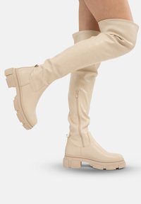 Botas de cuero beige hasta la rodilla con suela gruesa, cierre lateral de cremallera y detalle de puño doblado. Textura suave y hardware minimalista.