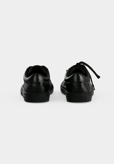 Paire de baskets en cuir noir vues de dos sur un fond blanc uni, avec un lacet défait.