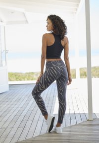 Zwarte crop tanktop gecombineerd met grijze leggings met een tijgerstreep patroon in beige. Model staat op een houten dek met witte sneakers.
