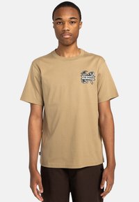 Tan bomulds T-shirt med en afslappet pasform, der har et sort grafisk tryk på venstre bryst og korte ærmer. Enkel rund halsudskæring.