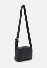 Bolso cruzado de piel negra con forma rectangular. Presenta un cierre con cremallera y una correa de tela con el logo "Calvin Klein". Textura suave.