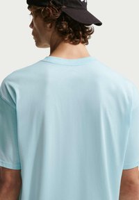 Νεαρός άνδρας με σγουρά μαλλιά, φοράει μαύρο καπέλο με την κορυφή προς τα πίσω και ανοιχτό μπλε μπλουζάκι, κοιτάζει προς τα πίσω με φόντο έναν απλό τοίχο.