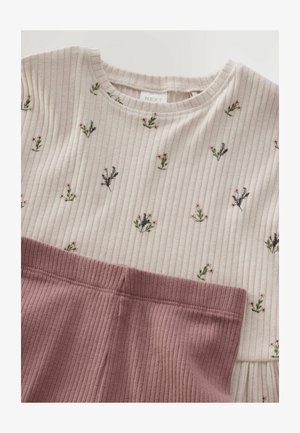 Rebrasti ružičasti top s cvjetnim aplicima u zelenoj i svijetloružičastoj boji, u kombinaciji s jednobojnim svijetloružičastim rebrastim hlačama. Mekana tekstura i prilagođeni dizajn.