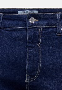 Jeans de mezclilla azul oscuro con costuras beige visibles, botón metálico y etiqueta que dice "ONLY & SONS" en la cinturilla.