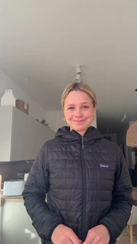 Femeie zâmbitoare cu păr blond poartă o jachetă neagră Patagonia, stând într-o bucătărie interioară cu dulapuri, prăjitor de pâine și corp de iluminat vizibile.