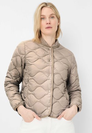 Frau mit blondem Haar, die eine taupefarbene gesteppte Jacke mit Druckknöpfen trägt, Hände in den Taschen, stehend vor einem einfarbigen hellen Hintergrund.