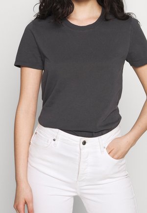 Vrouw draagt een effen donkergrijs T-shirt met korte mouwen, ingestopt in een witte jeans met hoge taille, één hand in de zak.