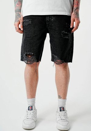 Jambes portant un short en jean noir déchiré, des chaussettes blanches, des baskets blanches, et des avant-bras tatoués avec les mains détendues sur les côtés.