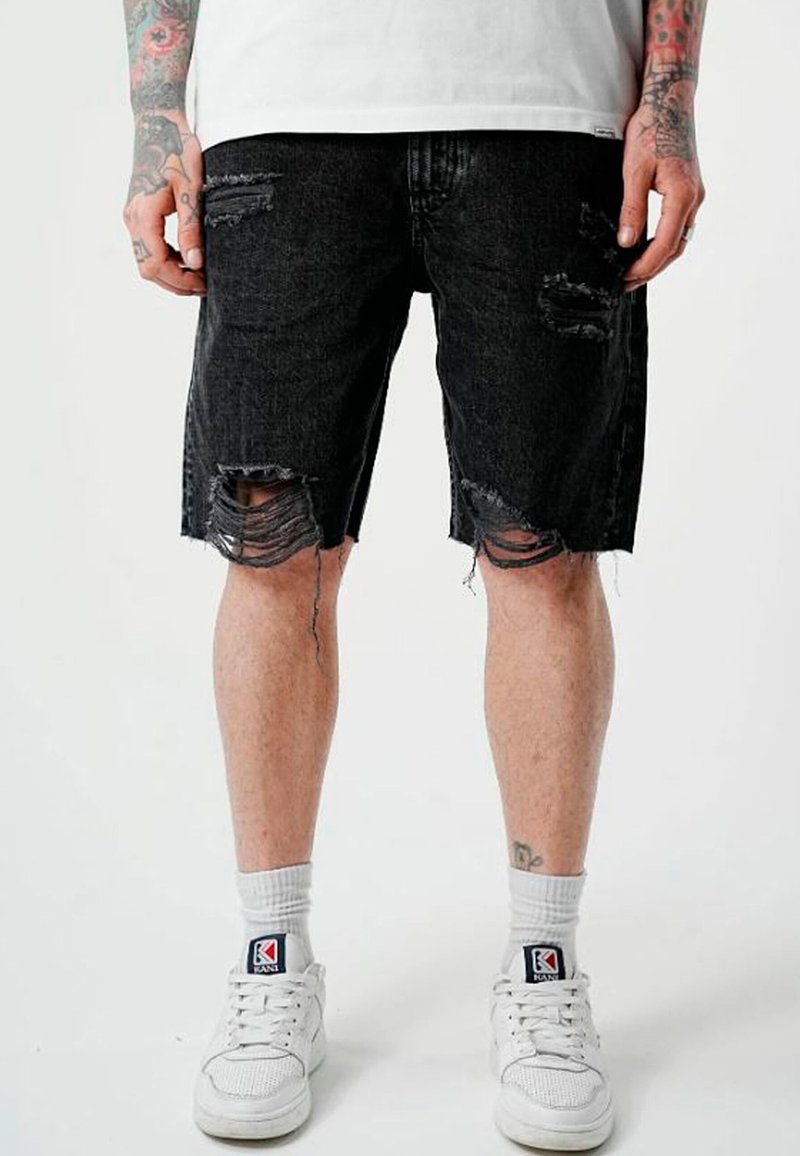 Jambes portant un short en jean noir déchiré, des chaussettes blanches, des baskets blanches, et des avant-bras tatoués avec les mains détendues sur les côtés.