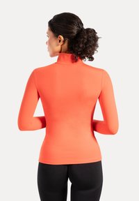 Strakzittend, langärmelig top in feloranje, met een hoge kraag en een gladde, rekbare stof. Draagt met zwarte leggings.